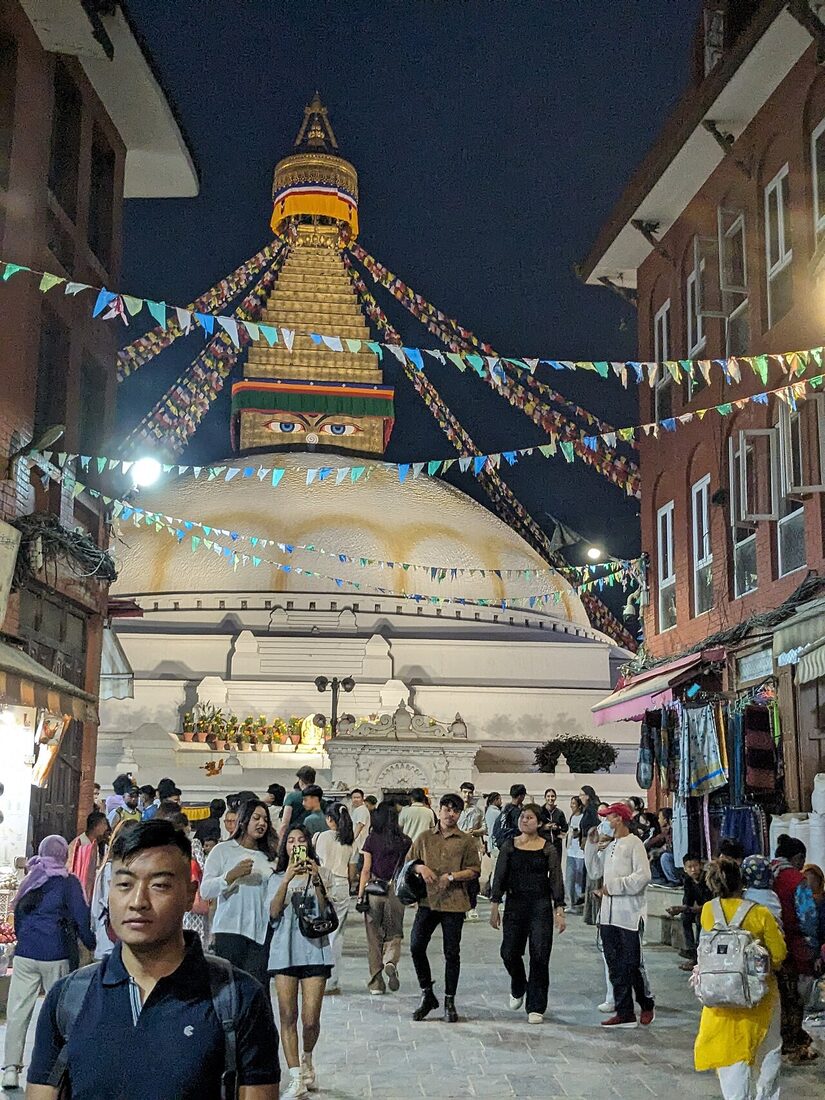 The lane into Boudha at dusk seen from the main gate: prayer flags strung overhead, lit shopfronts either side, people walking in toward the stupa
