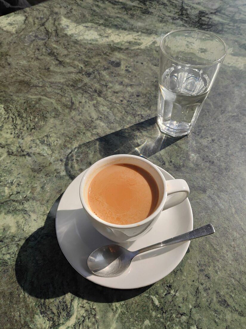A cup of milk tea on a saucer beside a glass of water, on a sunlit stone table