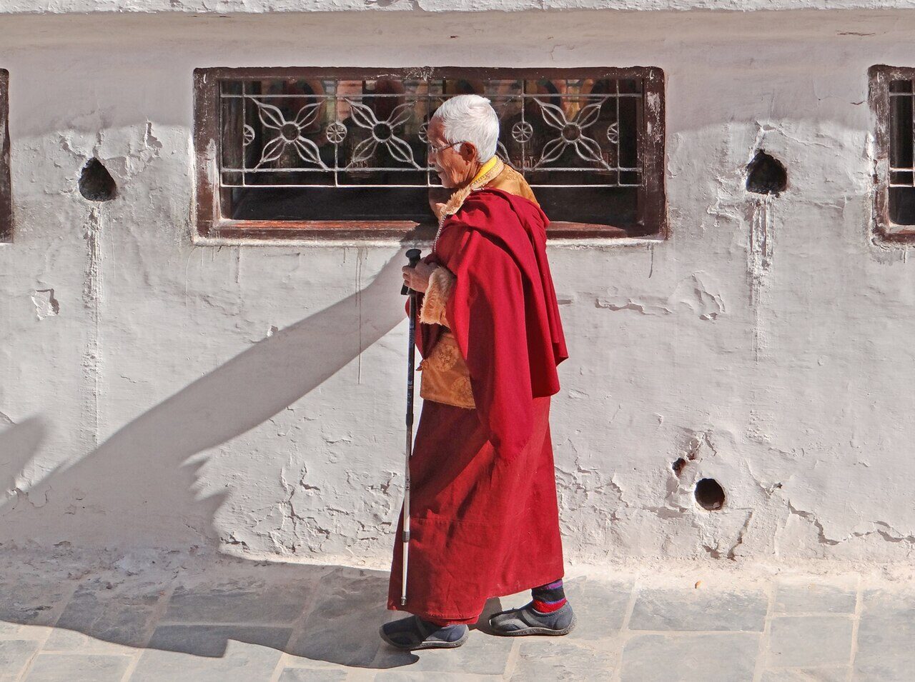 An elderly pilgrim in maroon robes, leaning on a stick, walking beside the whitewashed wall of the stupa below a carved window
