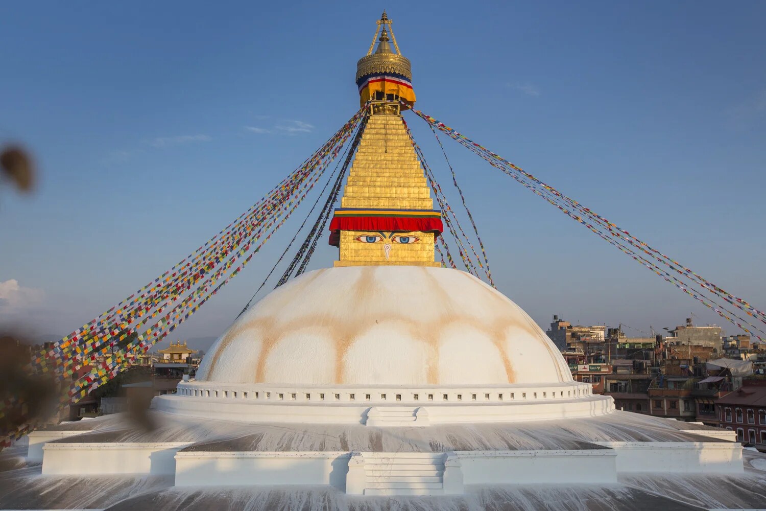 The gilded spire of the Great Boudhanath Stupa against a clear sky, the painted eyes of the Buddha above the whitewashed dome and prayer flags running out from the tower